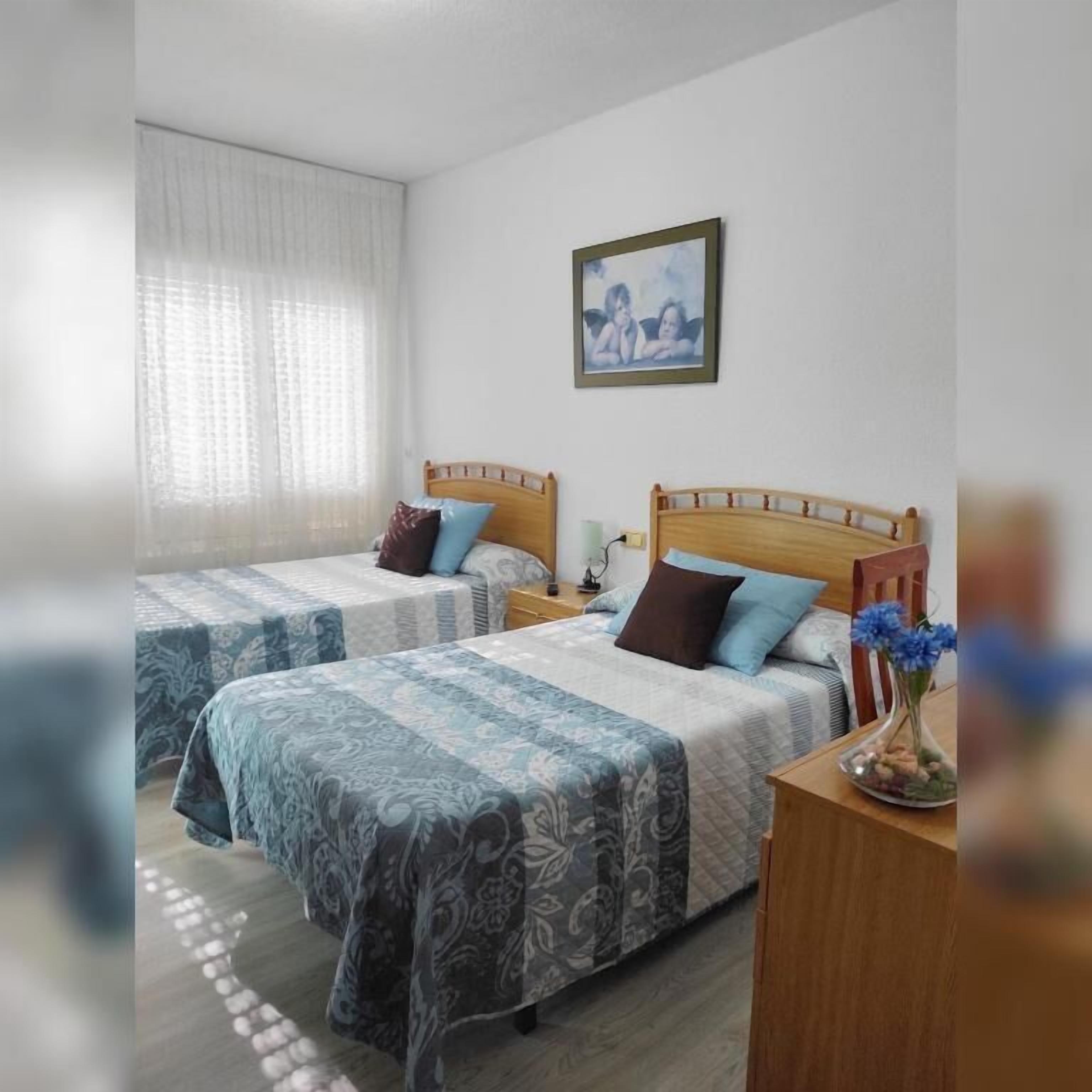 Habitación de hotel con dos camas, una mesita de noche con lámpara, un cuadro en la pared y un florero con flores en la mesita de noche.