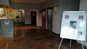 Un hall d’hôtel comprenant un comptoir d’accueil, un tableau blanc affichant des consignes de sécurité et un ascenseur.