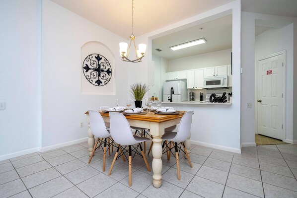 Un coin repas avec une table en bois, des chaises blanches, un lustre et une cuisine en arrière-plan.