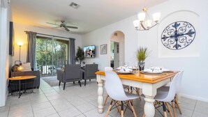 A dining area with a wooden table, white chairs, and a chandelier. There is a TV mounted on the wall and a ceiling fan. - Dream Cozy 3BR Condo - Disney 5 Min, Walk to Pool! (Orlando)