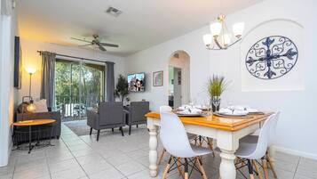 A dining area with a wooden table, white chairs, and a chandelier. There is a TV mounted on the wall and a ceiling fan.