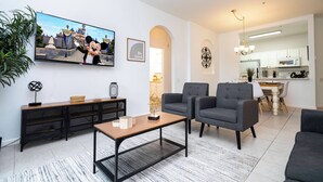 A modern living room with a flat-screen TV, grey armchairs, a coffee table, and a dining area with a kitchen in the background. - Dream Cozy 3BR Condo - Disney 5 Min, Walk to Pool! (Orlando)