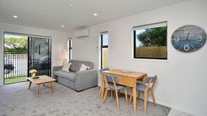 A modern living room with a grey sofa, wooden coffee table, and chairs. A large wall clock and a window with a view of greenery are visible. - Sea Air and Sandy Beach Beautiful 1 bed 1 bath (Christchurch)