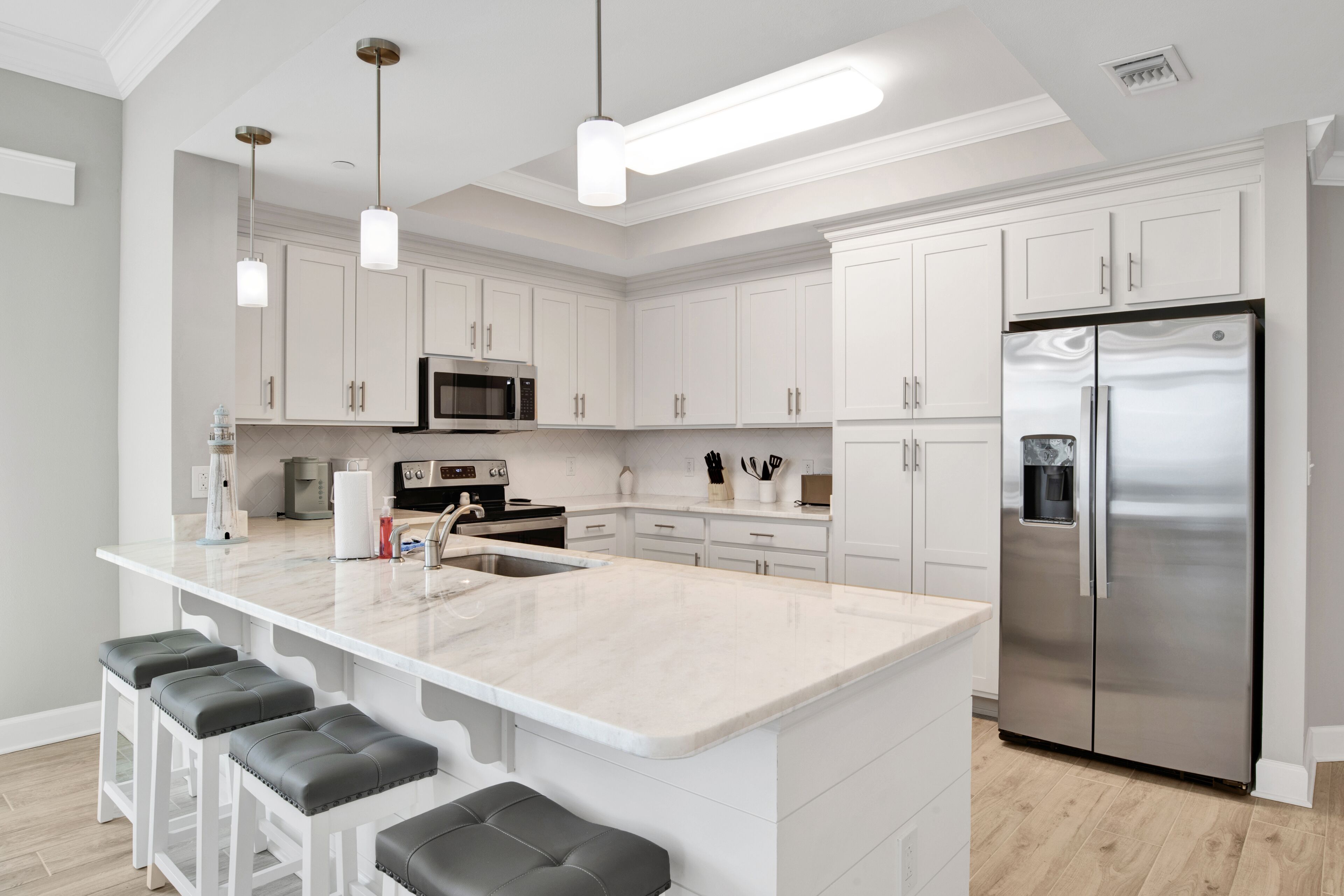 A modern kitchen with white cabinetry, a stainless steel refrigerator, a large island with bar stools, and pendant lighting.