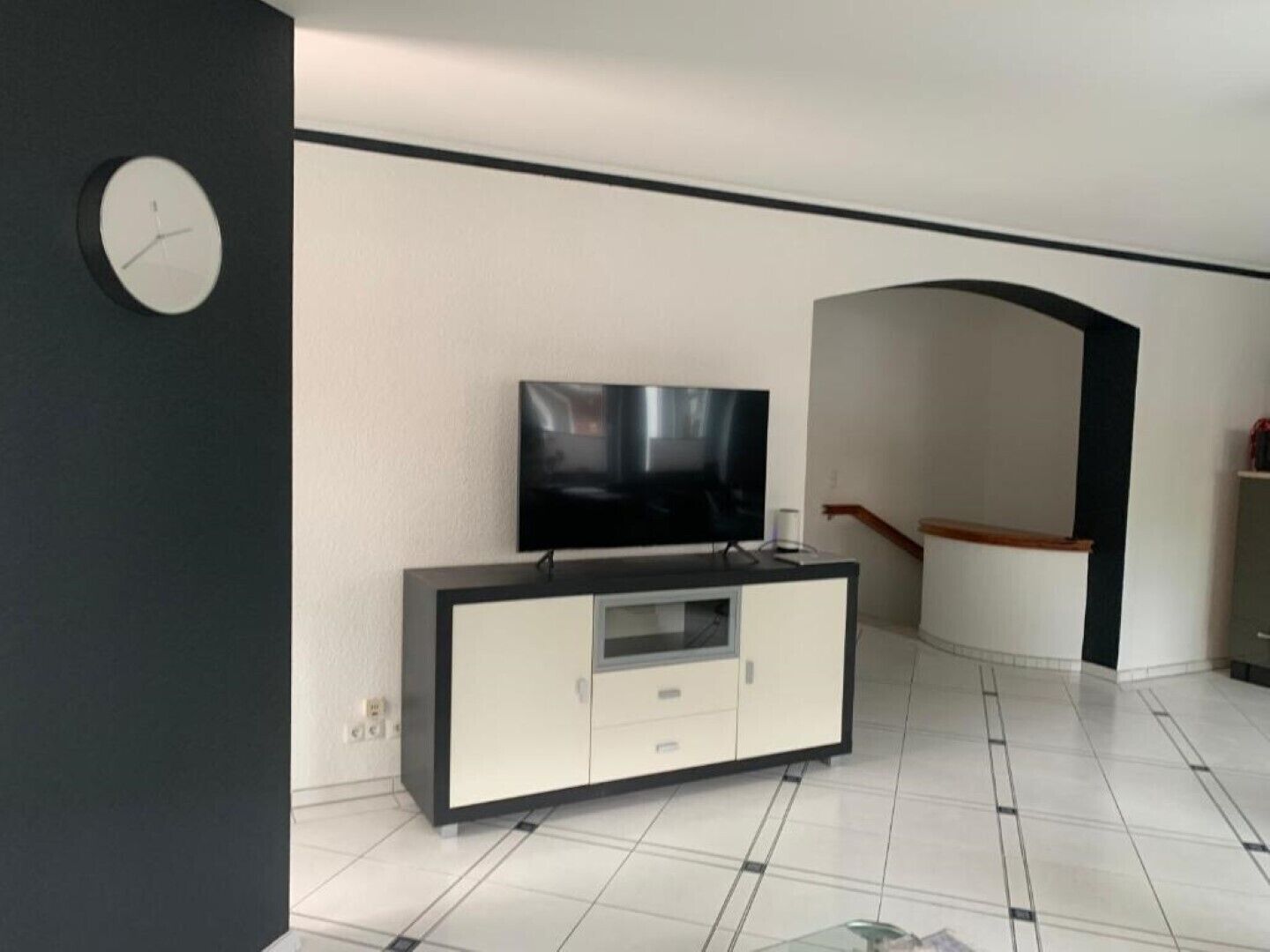 A modern living room with a flat-screen TV on a black and white cabinet, a clock on the wall, and a staircase leading to another level.