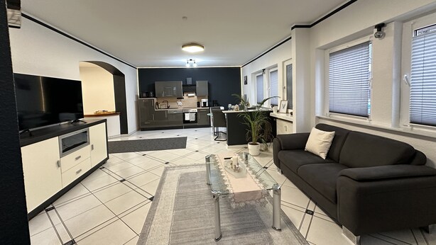 Een moderne woonkamer met een zwarte bank, een glazen salontafel en een keuken op de achtergrond.