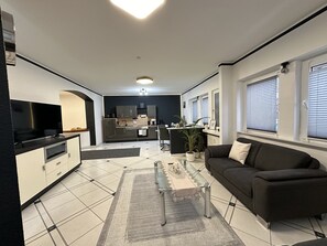 A modern living room with a black sofa, a glass coffee table, and a kitchen in the background.