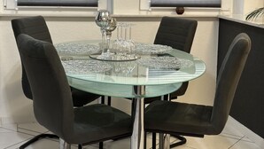 Een eetruimte met een glazen tafel omringd door vier zwarte stoelen, twee ramen met zonwering, en een beveiligingscamera boven de ramen.
