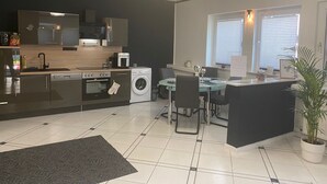 Een moderne keuken met een vaatwasser, oven en magnetron. Er is een eethoek met een tafel en stoelen. In de hoek staat een potplant.