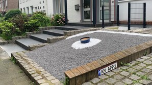 Een woongebouw met een verharde oprit, een grindgebied met een cirkelvormig patroon, en een houten plantenbak met een bord waarop 'P-KM APP 1' staat.