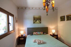 Habitación de hotel con una cama, dos mesitas de noche con lámparas, un cuadro en la pared y una ventana con vistas al follaje.