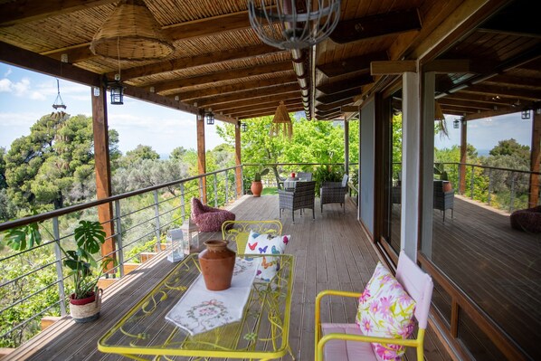 A wooden deck with yellow metal chairs and a table, surrounded by potted plants and a view of greenery.