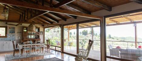 A spacious dining area with a wooden ceiling, large windows offering a view of the outdoors, and a cozy seating area with a rocking chair.