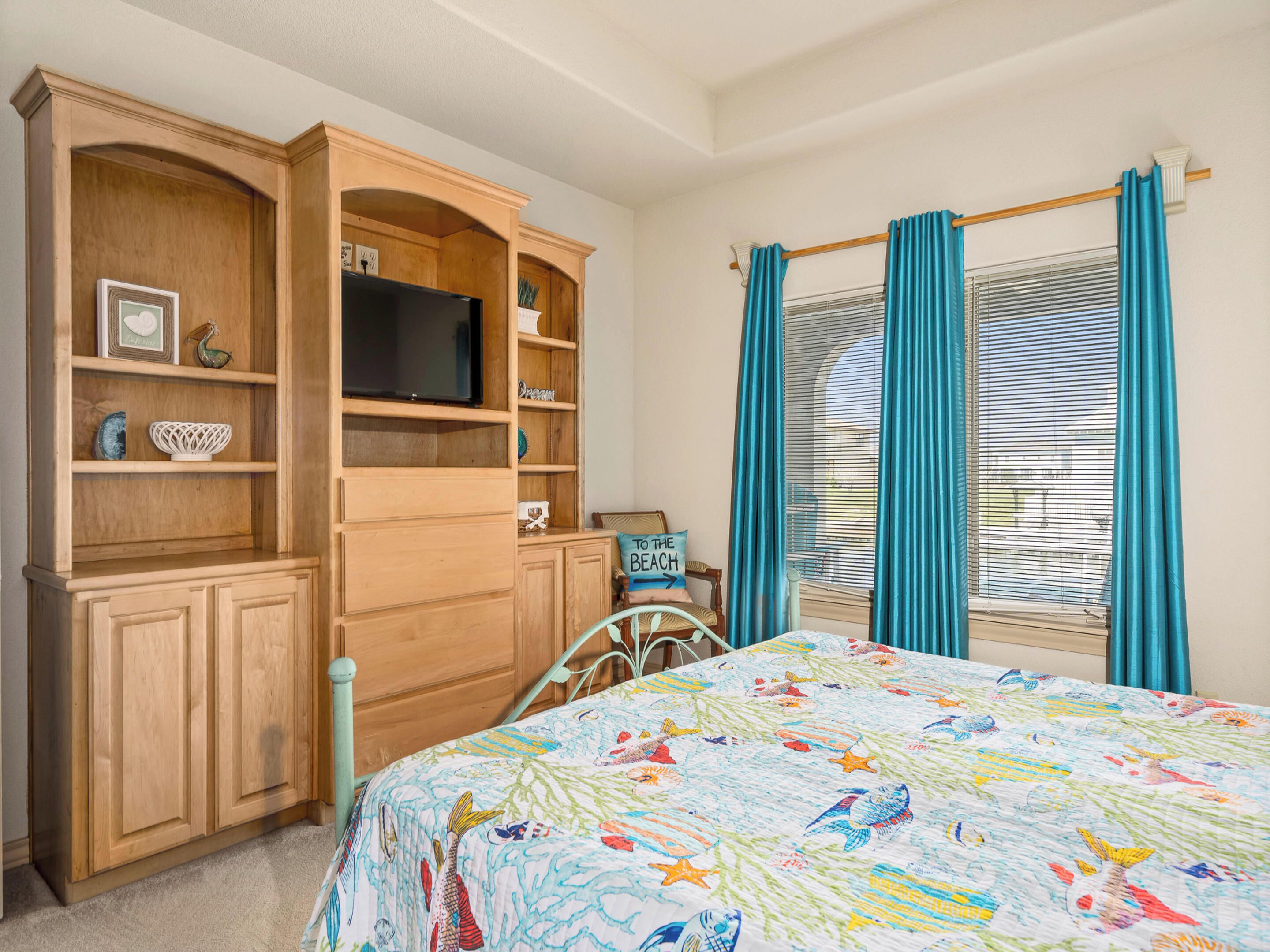 A bedroom with a bed, wooden built-in shelving, a television, and a window with curtains.