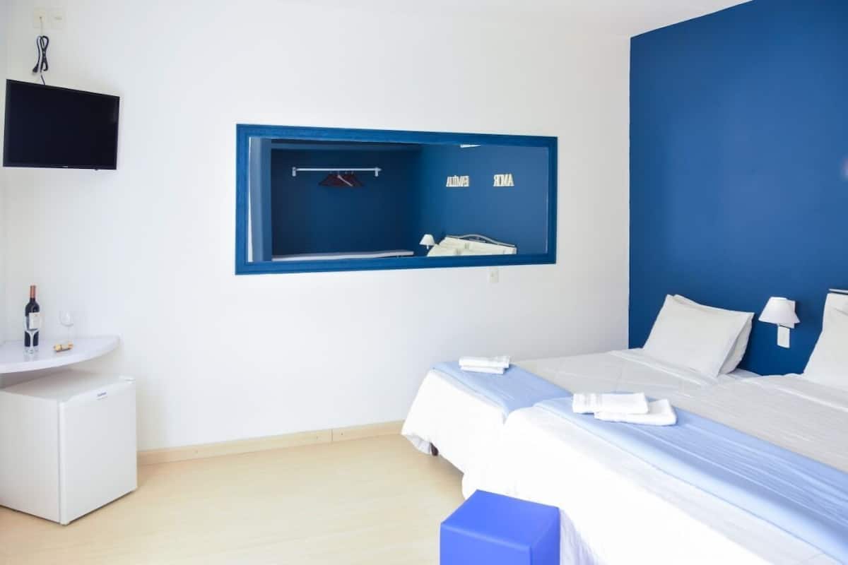 Ein Hotelzimmer mit einem Bett, einem kleinen Schreibtisch mit Flasche und Gläsern, einem an der Wand montierten Fernseher und einem spiegel mit blauem Rahmen.