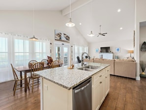 A spacious kitchen with a central island, wooden dining table, and chairs. There is a built-in dishwasher and a ceiling fan. - Port Royal Dream by Miss Kitty’s (Aransas Pass)