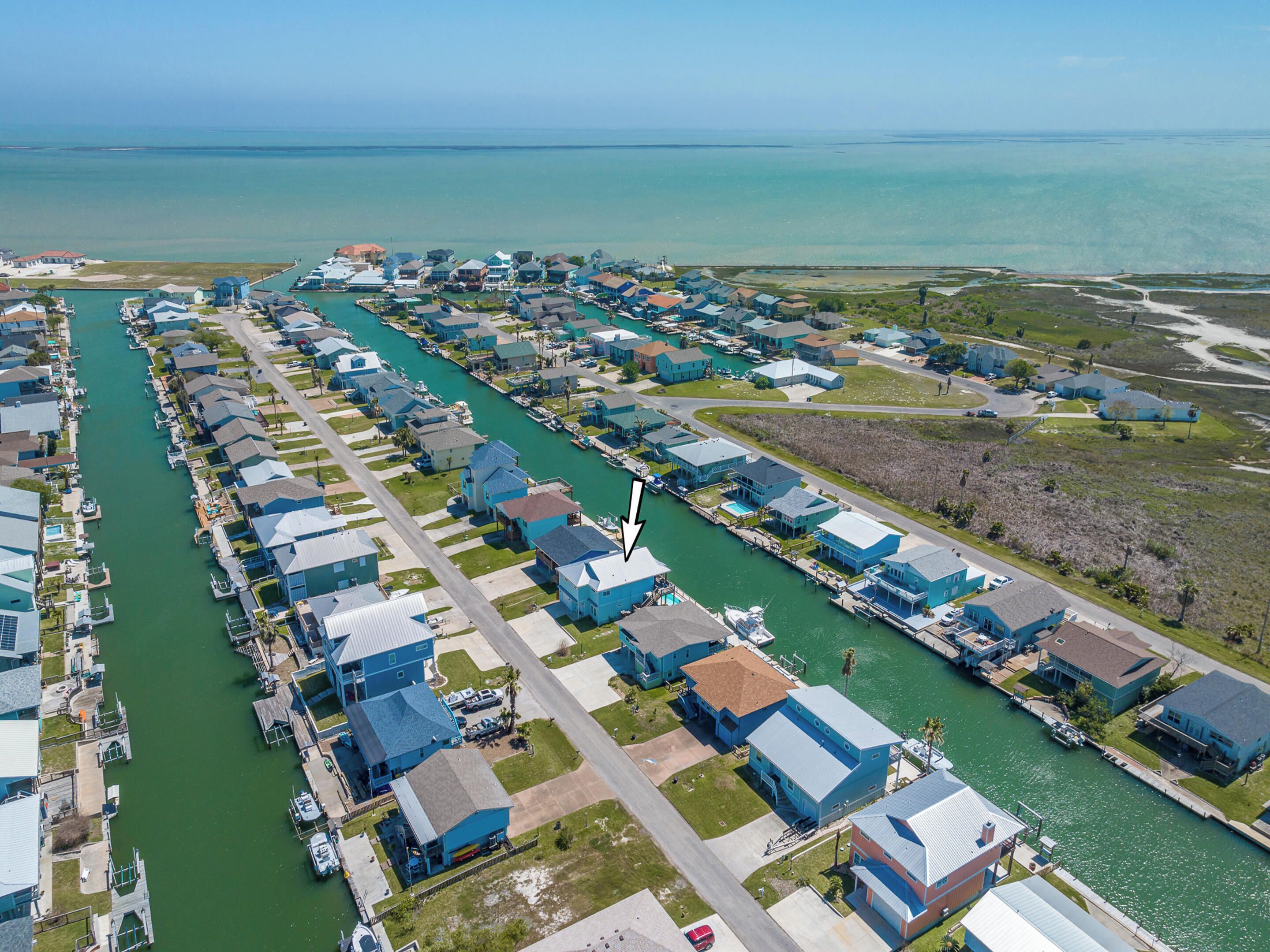 Una zona residencial costera con casas alineadas a lo largo de un canal estrecho, barcos amarrados en el puerto deportivo y una vista despejada del océano.
