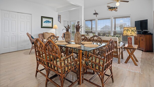 A spacious dining area with a glass-top table surrounded by wicker chairs, a ceiling fan, and a television in the background.