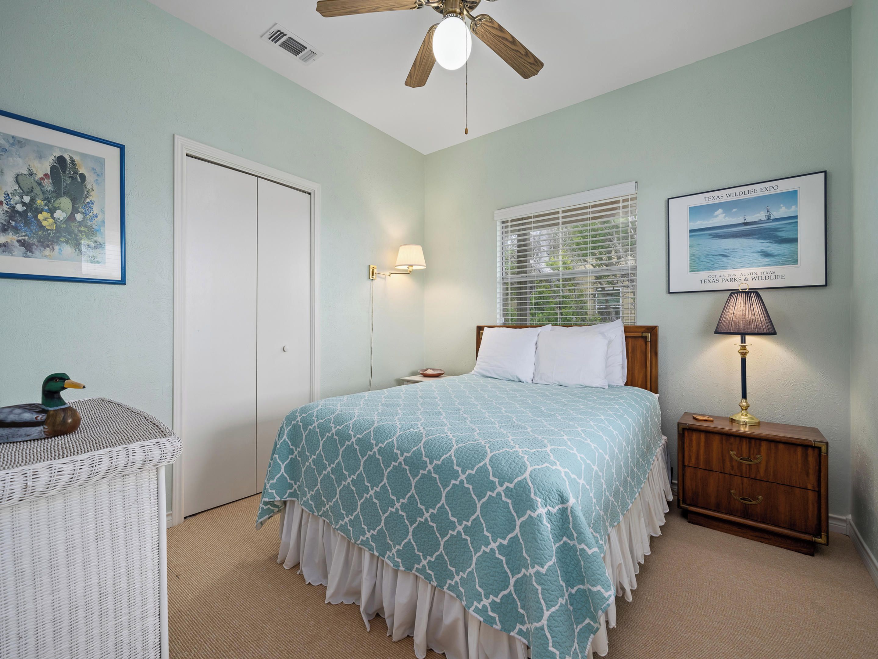 A bedroom with a bed, a nightstand, a lamp, a wicker chair, and a framed picture on the wall.