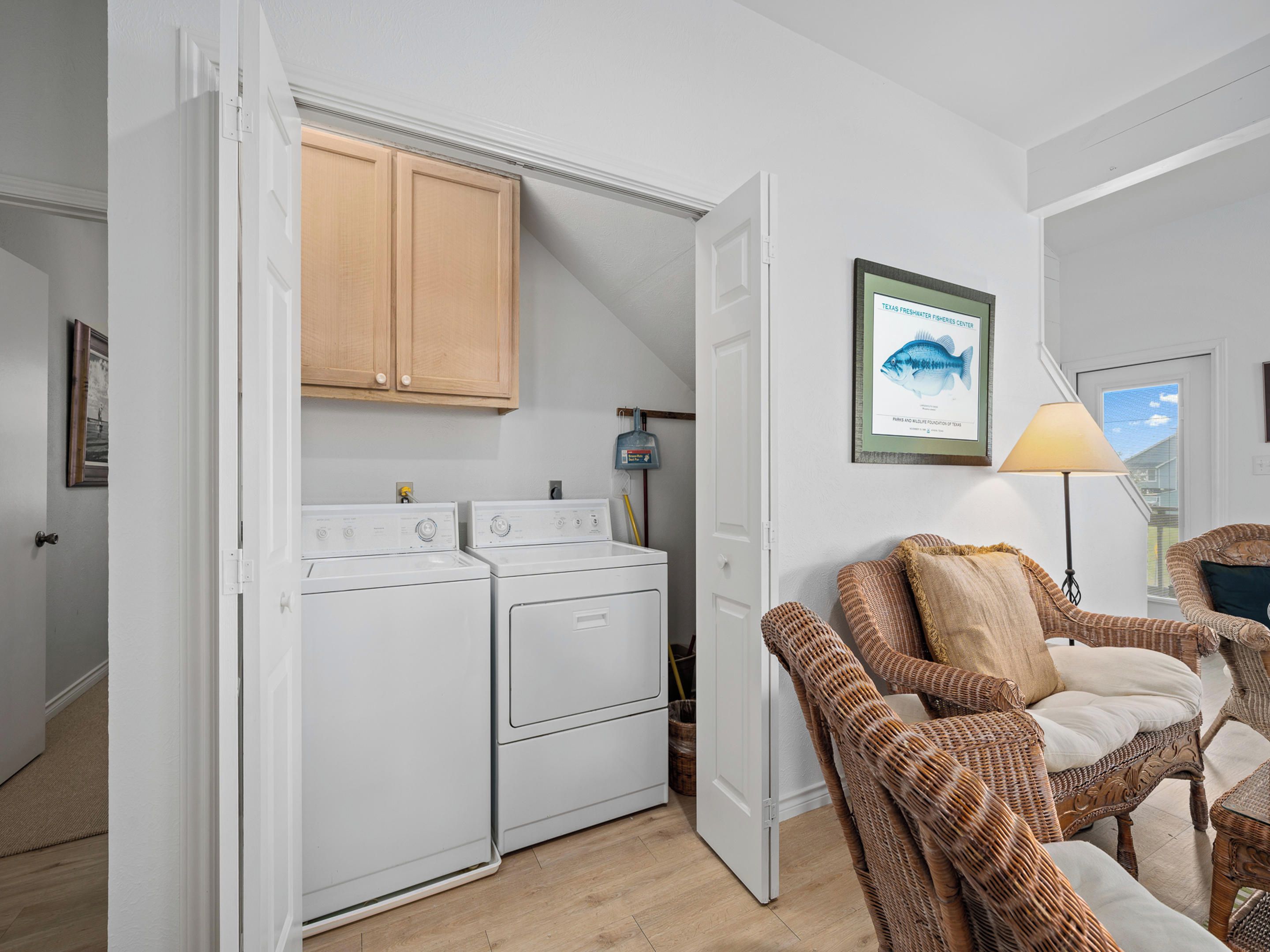 A compact kitchen with two white appliances, wooden cabinets, and a wicker chair in a bright room with a view.