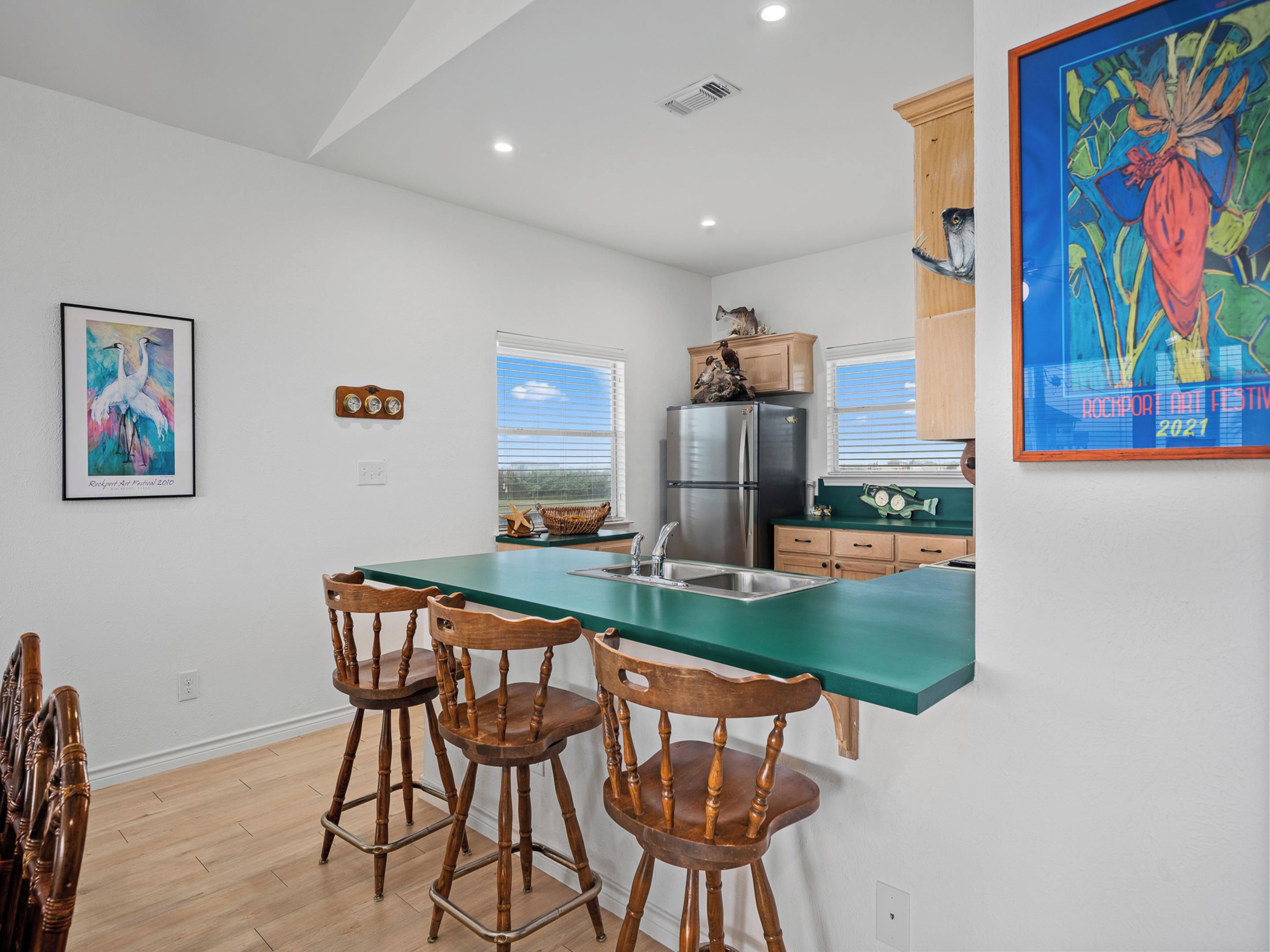 A modern kitchen with a green countertop, wooden chairs, a stainless steel refrigerator, and a painting on the wall.