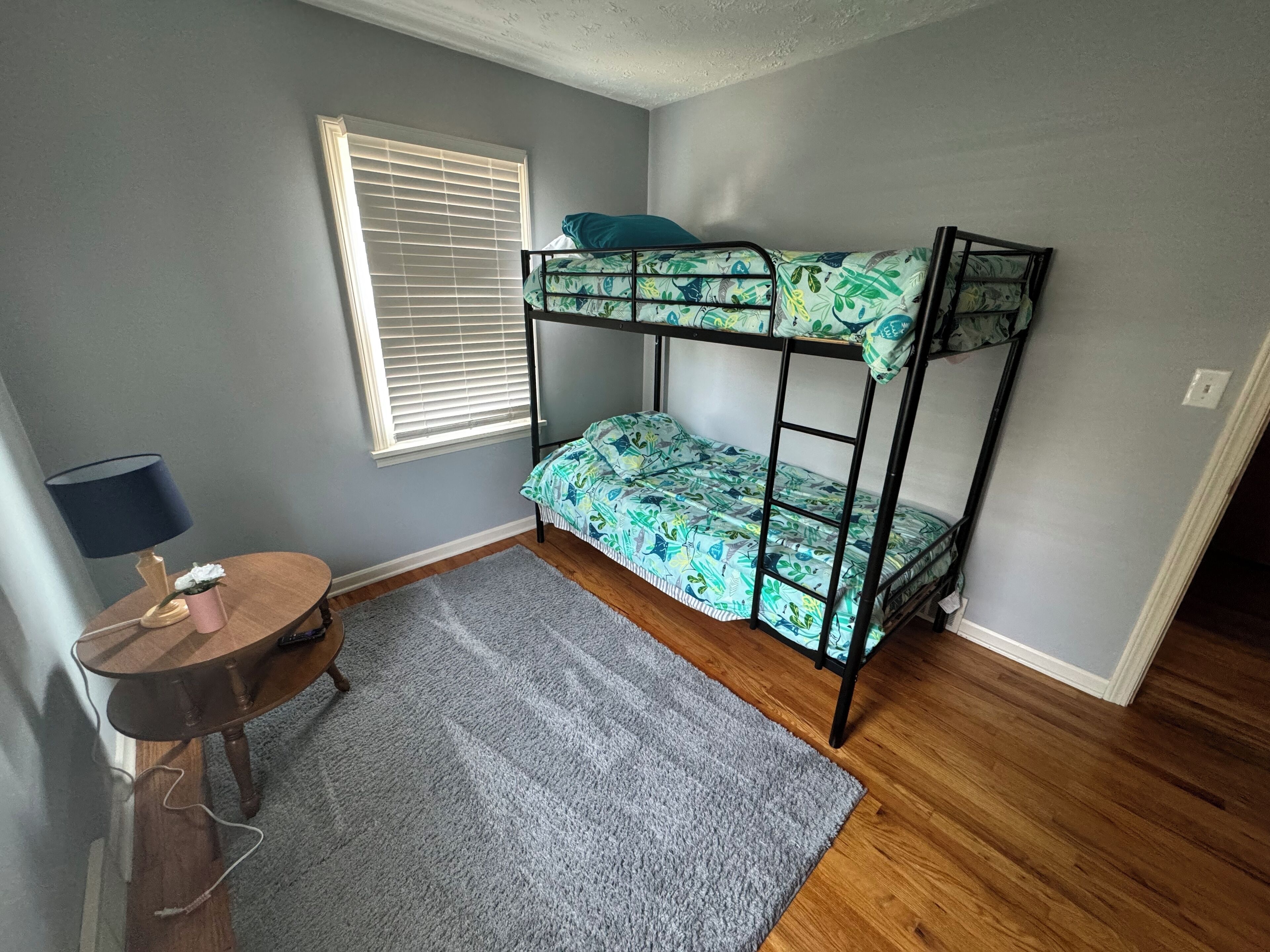 A small room with a bunk bed, a desk with a lamp, a window with blinds, and a grey rug.