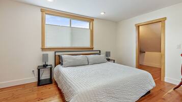 Un dormitorio con una cama grande, una ventana con persianas, una mesita de noche con lámpara y suelo de madera.