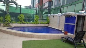 Une piscine dont le bord est recouvert de carreaux, entourée d’un petit muret et d’une petite pelouse où se trouve une chaise.