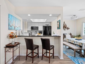 A modern kitchen with white cabinets, a black refrigerator, and a microwave. There is a dining area with a table and chairs, and a vase of flowers on a side table. - Gaetano's Getaway by Miss Kitty’s (Rockport)