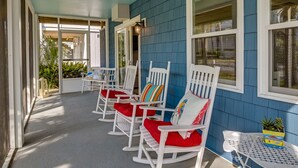 A covered porch with white rocking chairs and red cushions, a small table with a plant, and a blue exterior wall. - 'Below Deck' Beach House: Walk to the Ocean! (Surfside Beach)