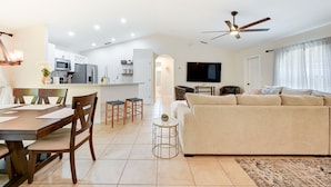 Uma sala de estar moderna com um sofá bege, mesa de jantar de madeira e cadeiras. É visível uma cozinha com armários e eletrodomésticos brancos ao fundo.