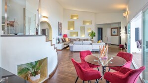 A spacious living area with a modern design, featuring a glass dining table, red upholstered chairs, and a white sofa set.