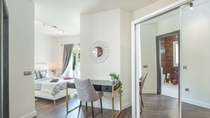 Ein modernes Schlafzimmer mit Bett, Nachttisch und Schreibtisch mit Stuhl. An der Wand befindet sich ein Spiegel und es gibt einen Blick in einen anderen Raum.