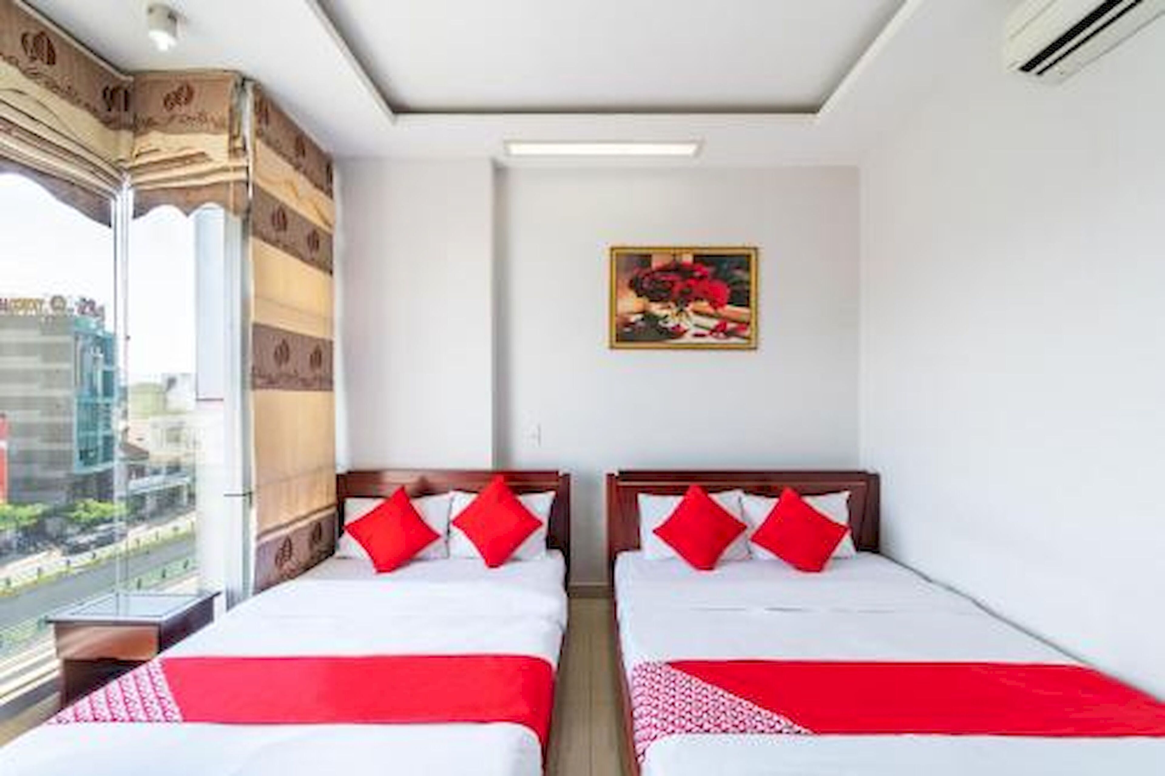 Hotelkamer met twee bedden, rode beddengoed, een schilderij aan de muur en uitzicht op de stad door grote ramen.