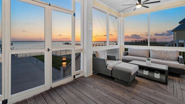 Eine überdachte Terrasse mit Blick auf den Sonnenuntergang über dem Wasser, Korbmöbel und ein Deckenventilator.