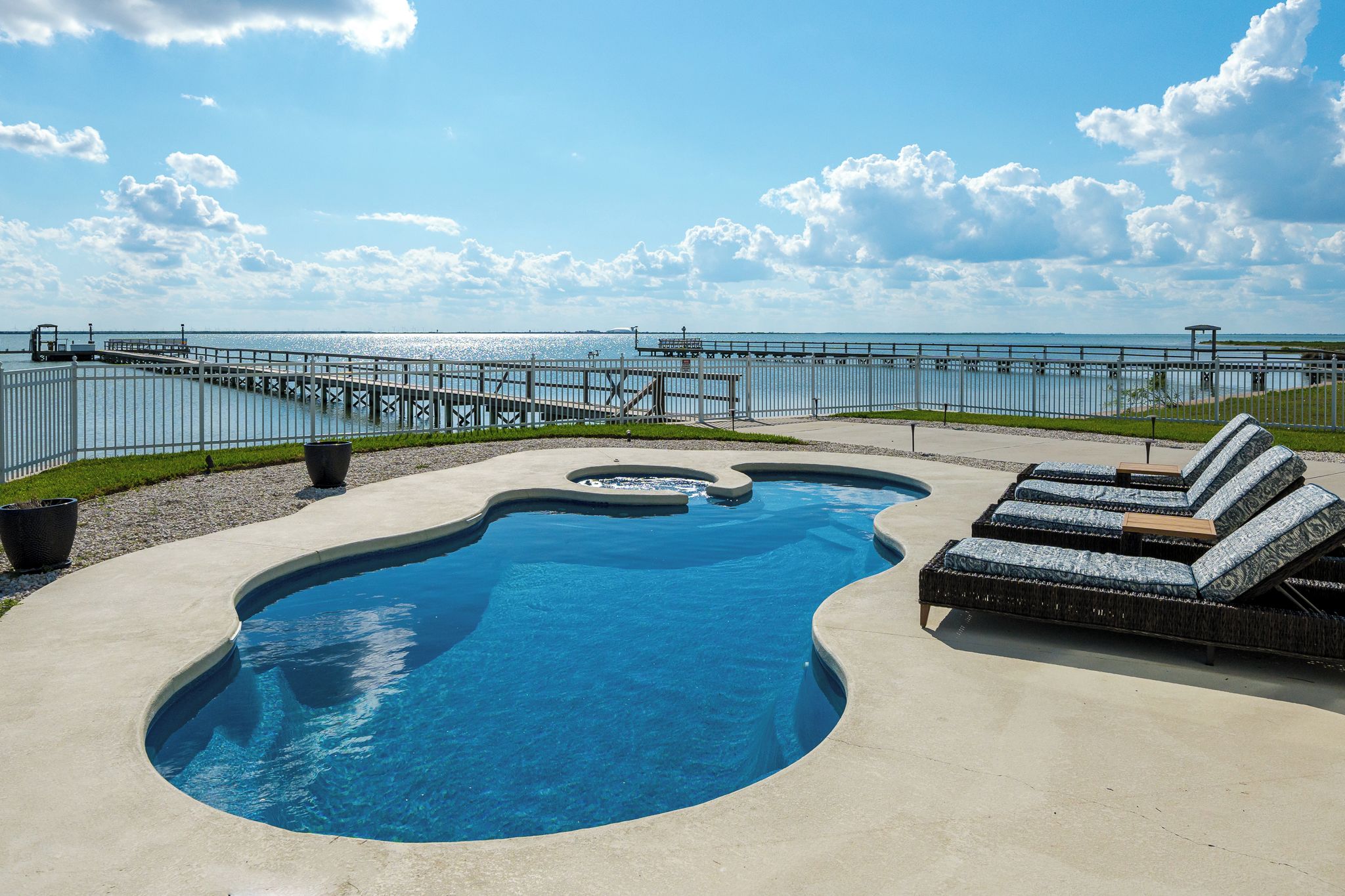 A swimming pool with a unique shape, surrounded by a concrete deck, a lounge area with cushions, and a view of the ocean and pier.