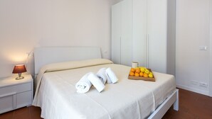 Una cama bien tendida con sábanas blancas, una lámpara de noche, una bandeja con naranjas y un armario blanco.