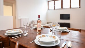 Una mesa de comedor dispuesta con platos, vasos, cubiertos, una botella de vino y un televisor al fondo.