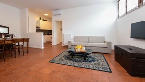 Una sala de estar con sofá, mesa de centro y televisor. Hay un comedor con mesa y sillas. El piso está cubierto de baldosas y tiene una alfombra grande.