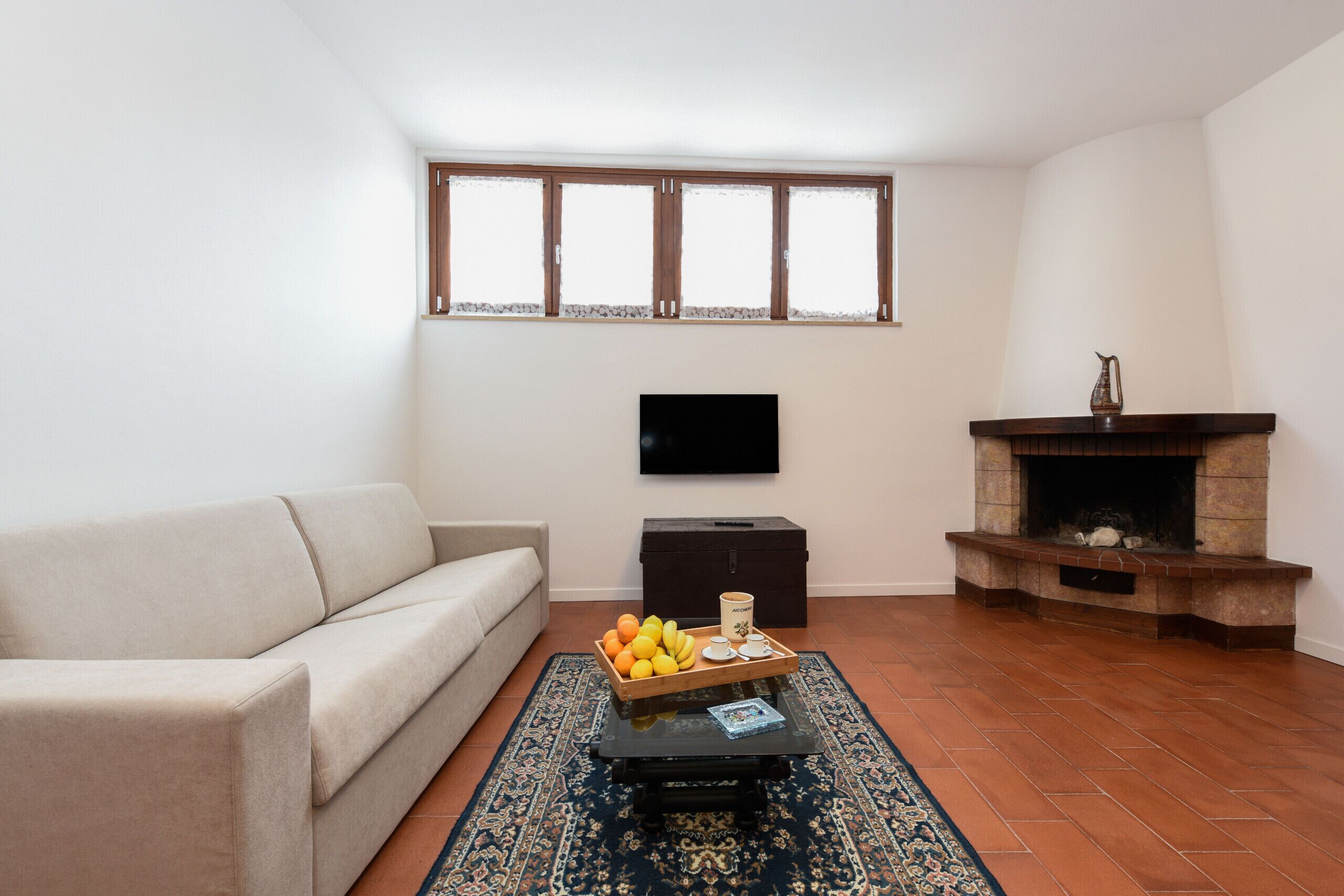 A living room with a sofa, a coffee table with fruit, a television, a fireplace, and a window with blinds.