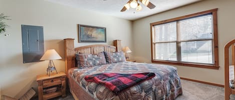 A bedroom with a bed, a ceiling fan, a window with blinds, a nightstand with a lamp, and a wall-mounted cabinet.