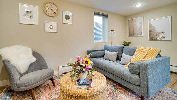 A living room with a grey armchair, a blue sofa, a round coffee table with a book and a vase of flowers, and a wall clock. - Alluring Anchorage Apt - Walk to Coastal Trl! (Anchorage)