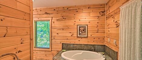 A wooden cabin bathroom with a large bathtub, a window with a view, and a framed picture on the wall.