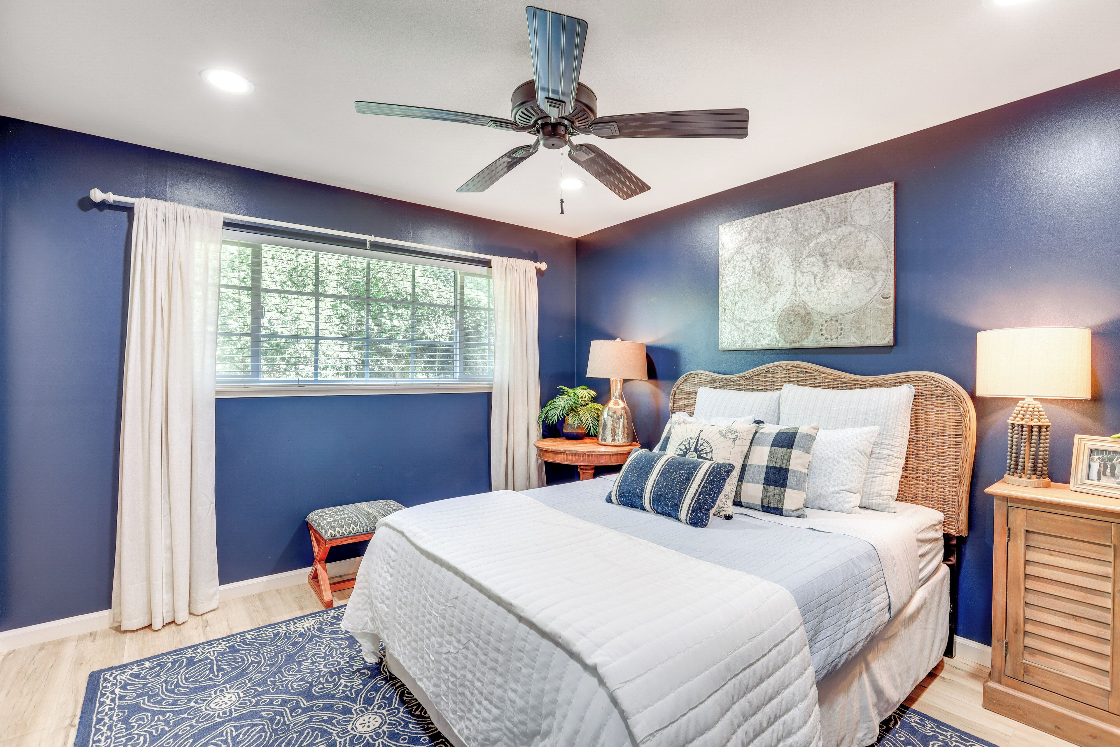 Une chambre à coucher comprenant un lit, un ventilateur de plafond, une fenêtre avec des rideaux, une table de chevet avec une lampe et une œuvre d’art accrochée au mur.