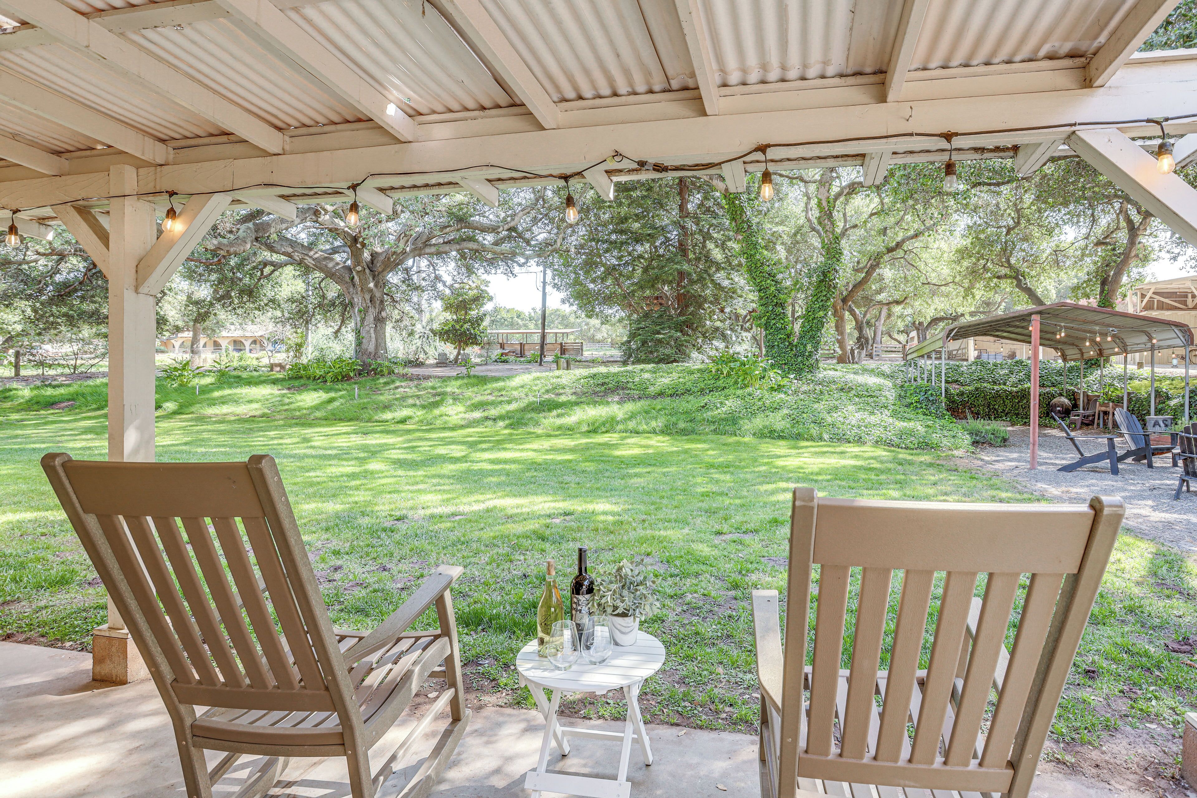 An outdoor patio with two wooden chairs, a small table with a bottle and glass, and a pergola with string lights.