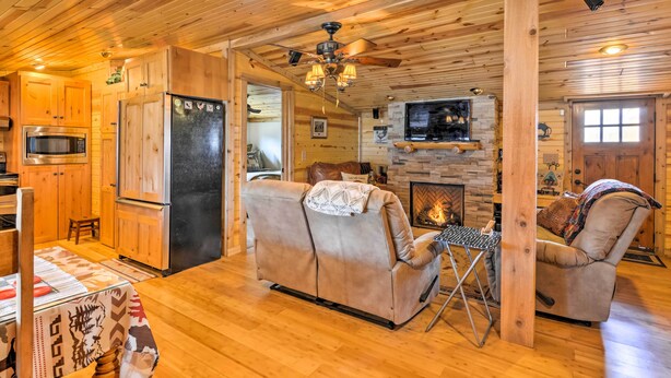 A cozy cabin interior with a wooden ceiling, a fireplace, a sofa, a dining table, and a kitchen area with a refrigerator and microwave.