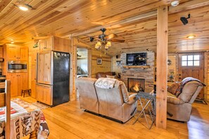 A cozy cabin interior with a wooden ceiling, a fireplace, a sofa, a dining table, and a kitchen area with a refrigerator and microwave.