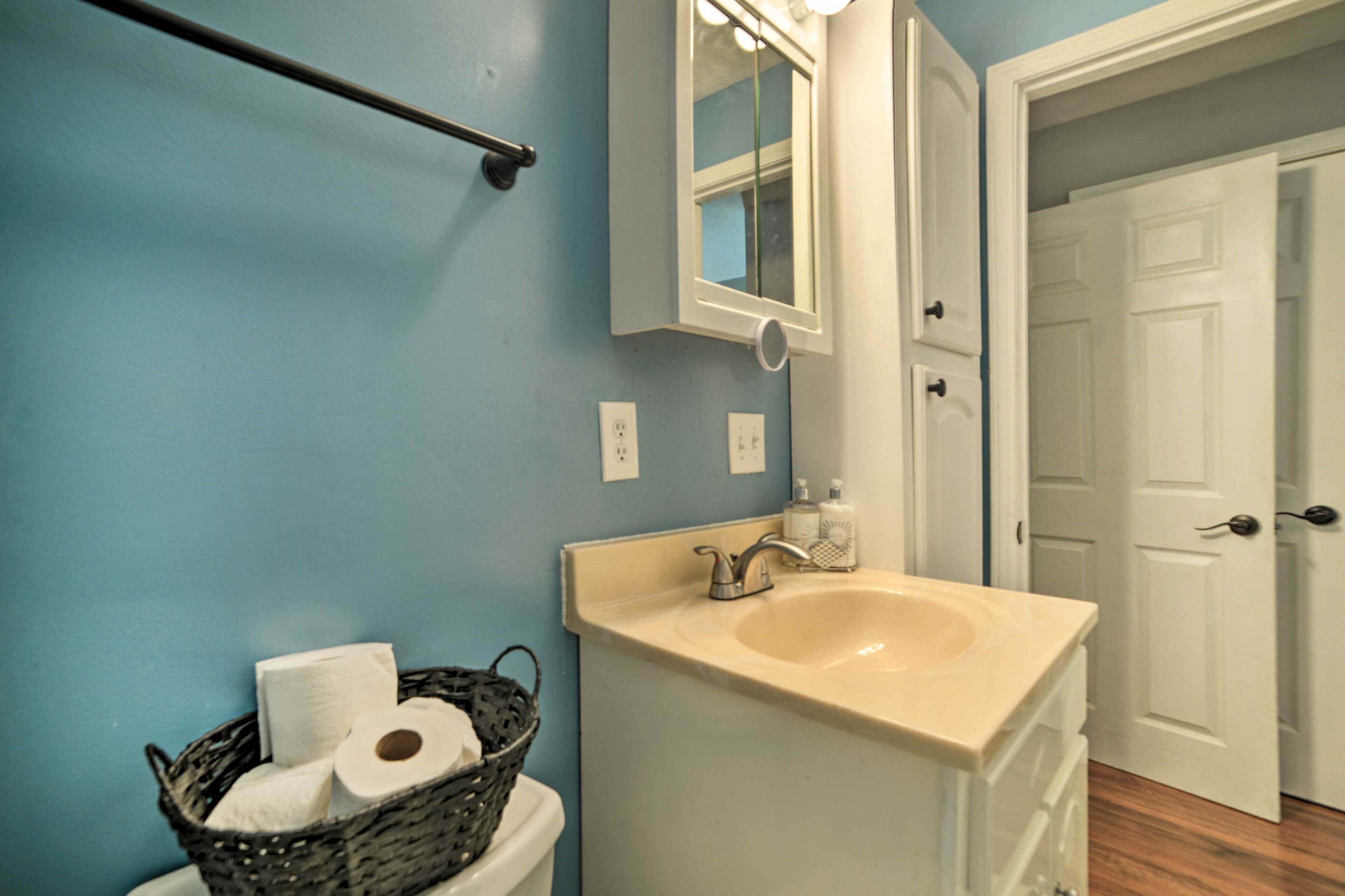A bathroom with a sink, mirror, and a basket of toilet paper on the wall.