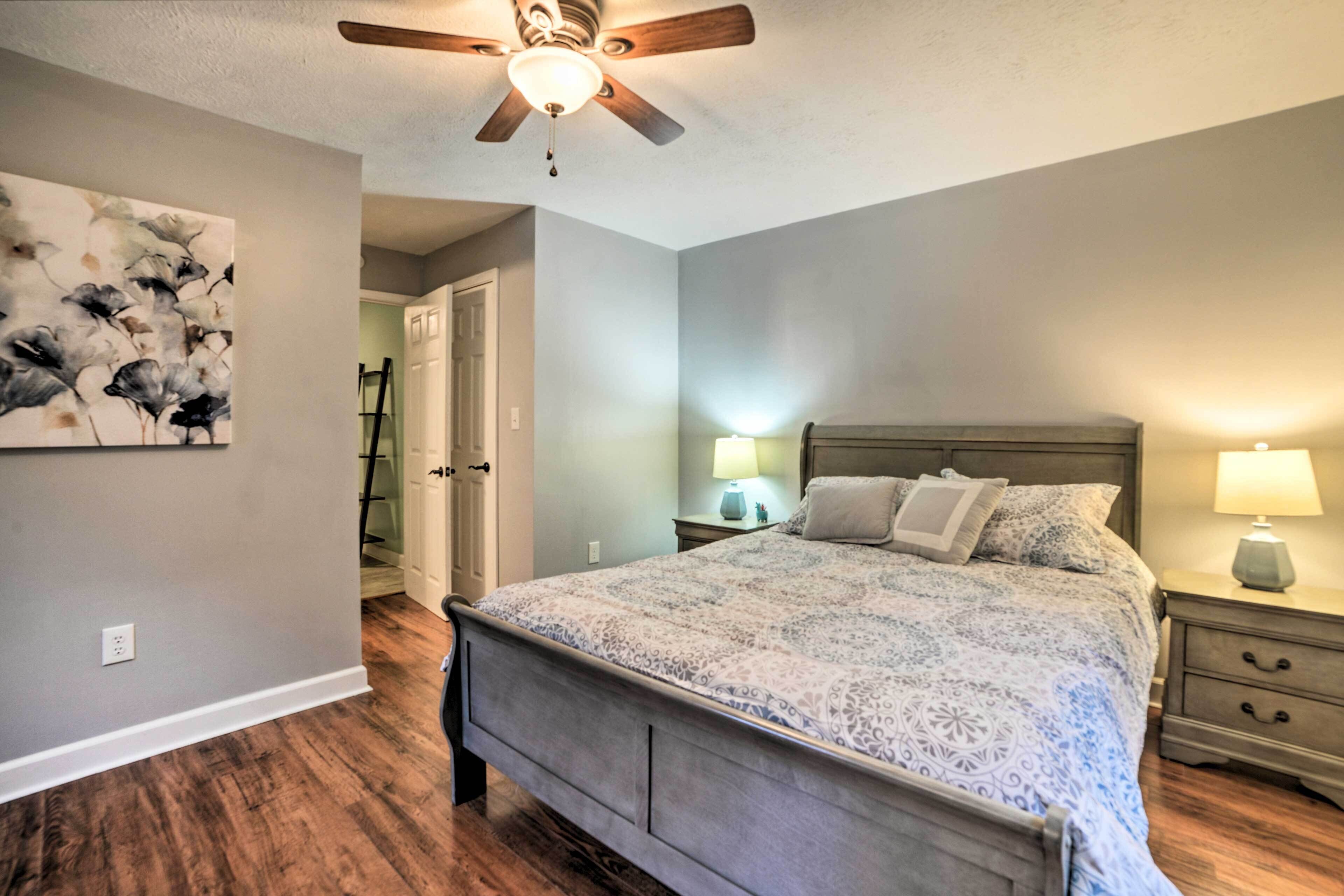 A bedroom with a bed, nightstands, a ceiling fan, a wall art piece, and a door leading to another room.