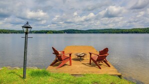 Eine hölzerne Anlegestelle mit zwei roten Adirondack-Stühlen und einer Feuerstelle, mit Blick auf einen stillen See mit bewaldeter Uferlinie unter einem bewölkten Himmel.